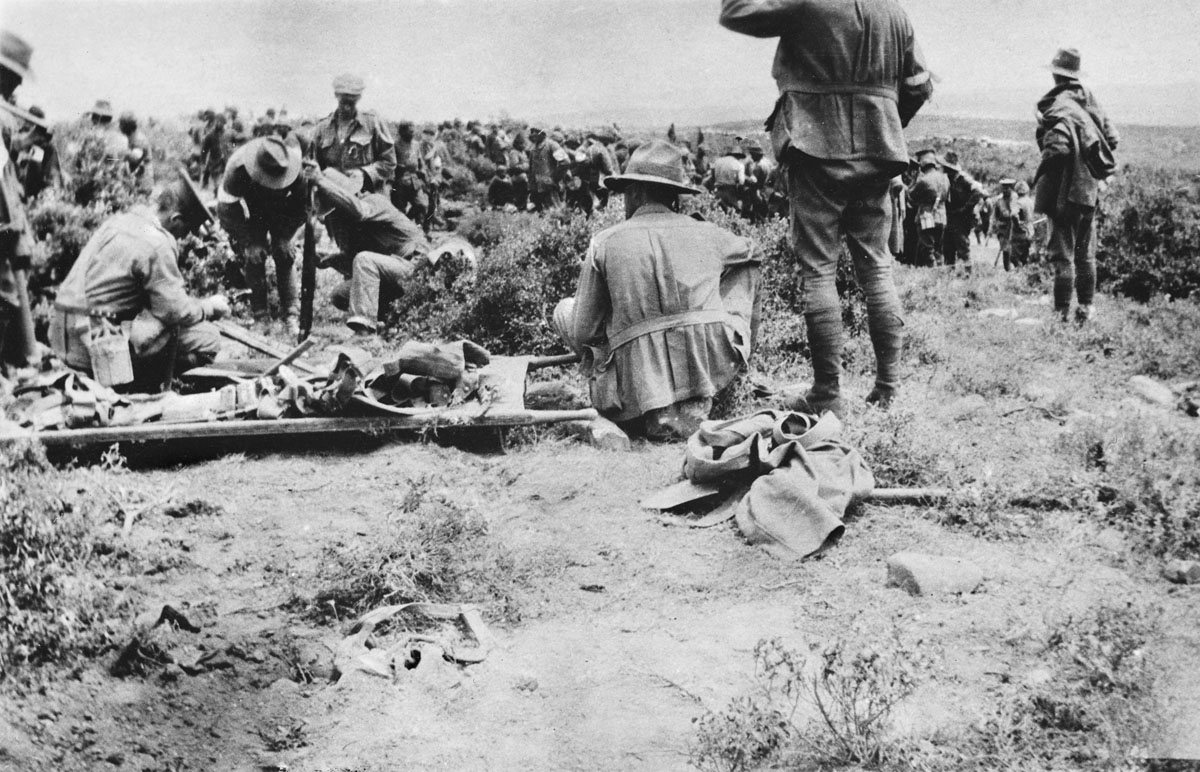 Australian troops collect equipment gathered from their dead, while in the background Turks bury several dozen of their troops.